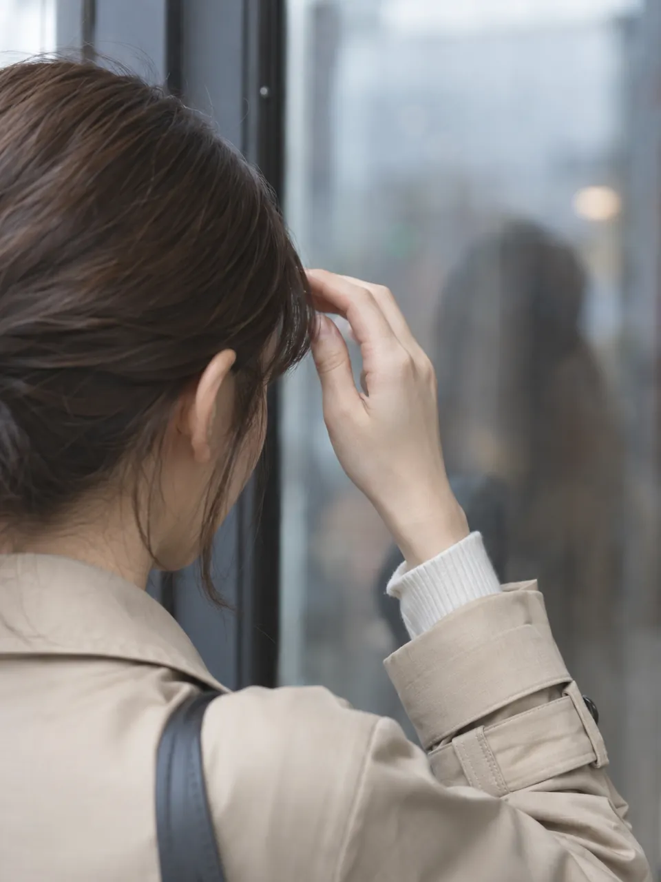 湿気の朝、ガラス戸の前で前髪を指で押さえる女性の後ろ姿のイメージ