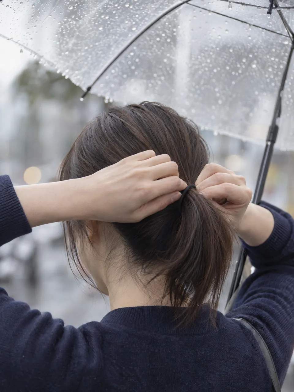 雨の日、傘をさしたまま髪をゴムで結ぶ女性の後ろ姿のイメージ